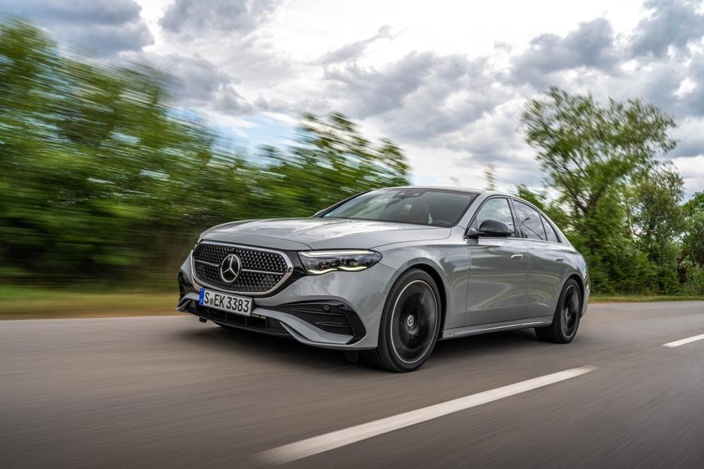 A silver-grey Mercedes-Benz E-Class Sedan driving on a country road with blurred trees behind it