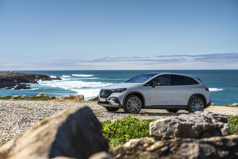 A light grey Mercedes-Benz EQE SUV parked on a pebble shore with the sea and rocks behind it