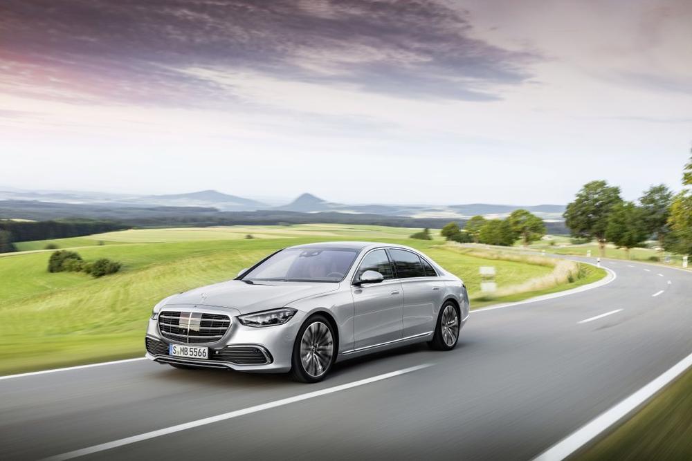 A silver Mercedes-Benz S-Class driving along a curving road through green hills under a soft evening sky